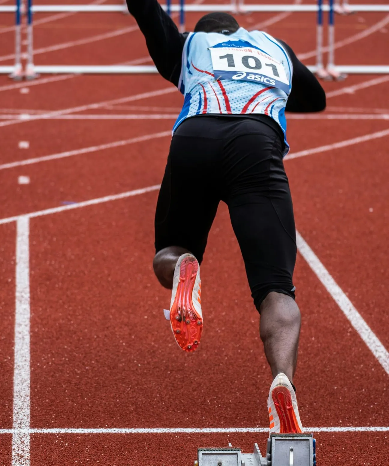 Sprinter seen from behind exploding out of the starting blocks on a red track, bib 101, hurdles ahead